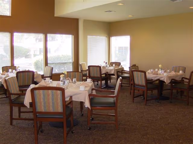 A dining room with several tables covered in light-colored tablecloths, each set with cups, glasses, and small flower vases. The room has large windows letting in natural light and wooden chairs with cushioned seats and backs arranged around the tables.