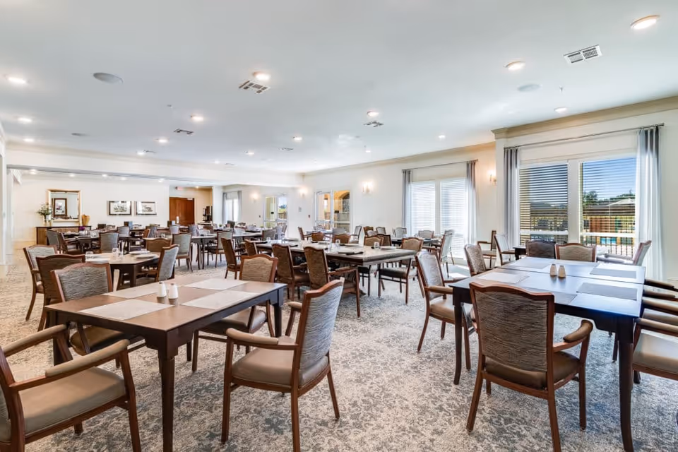 A spacious and well-lit dining room with multiple wooden tables and cushioned chairs arranged neatly. Large windows with curtains allow natural light to fill the room, and the carpeted floor features a subtle pattern. The walls are light-colored, and there are decorative mirrors and framed pictures on the far wall.