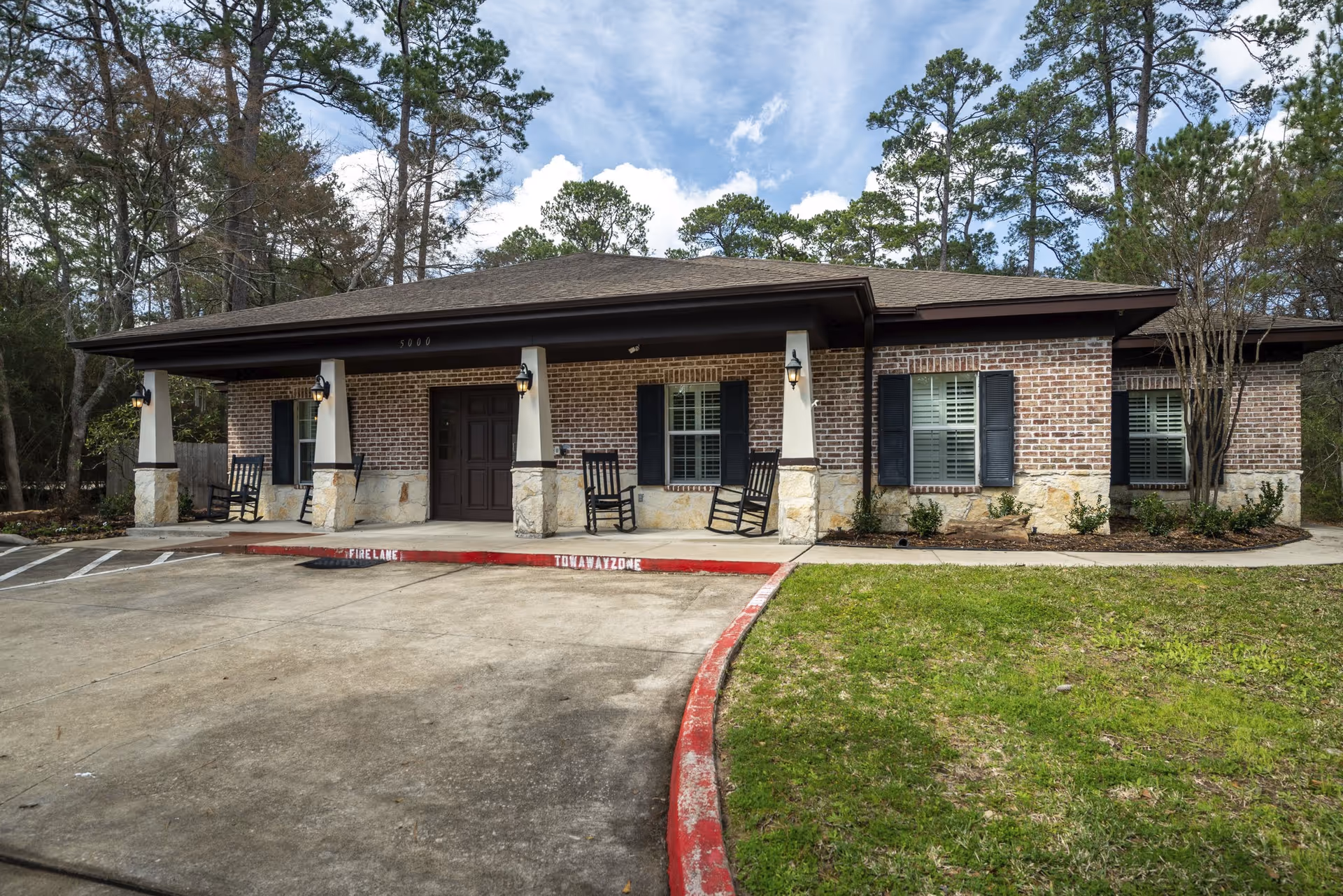 Exterior view of a single-story brick building with a covered porch supported by stone pillars. The porch has four black rocking chairs and wall-mounted lantern-style lights. The building is surrounded by trees and has a concrete driveway with a red-painted curb marked as a fire lane and tow away zone.