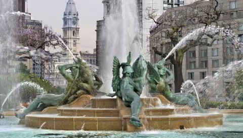 An ornate fountain sculpture in a public plaza with water jets and surrounding buildings.