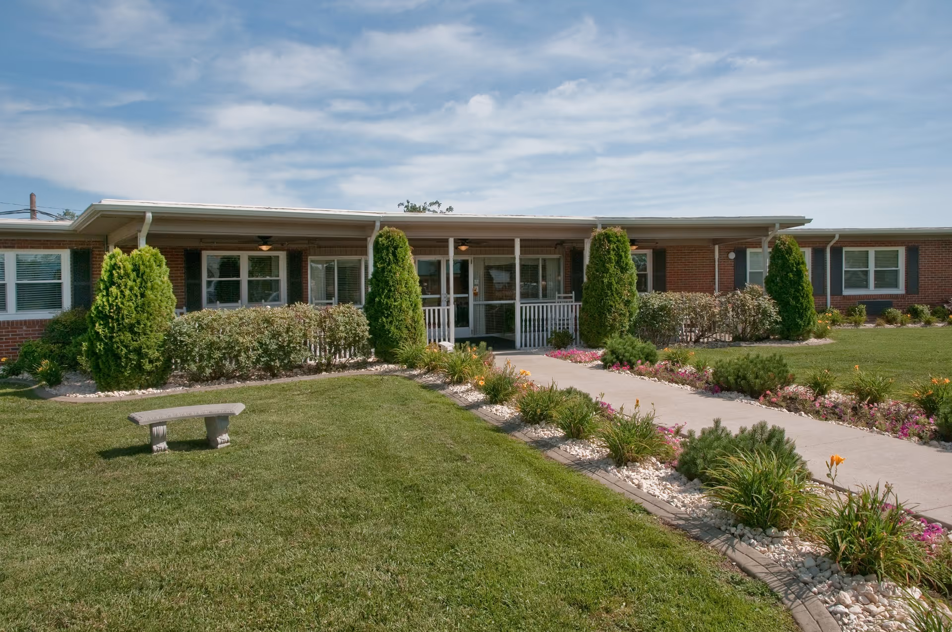 Front exterior view of a single-story brick building with a covered entrance, surrounded by neatly trimmed bushes, small trees, and a well-maintained lawn with a concrete walkway and flower beds on either side. A stone bench is placed on the grass near the walkway.