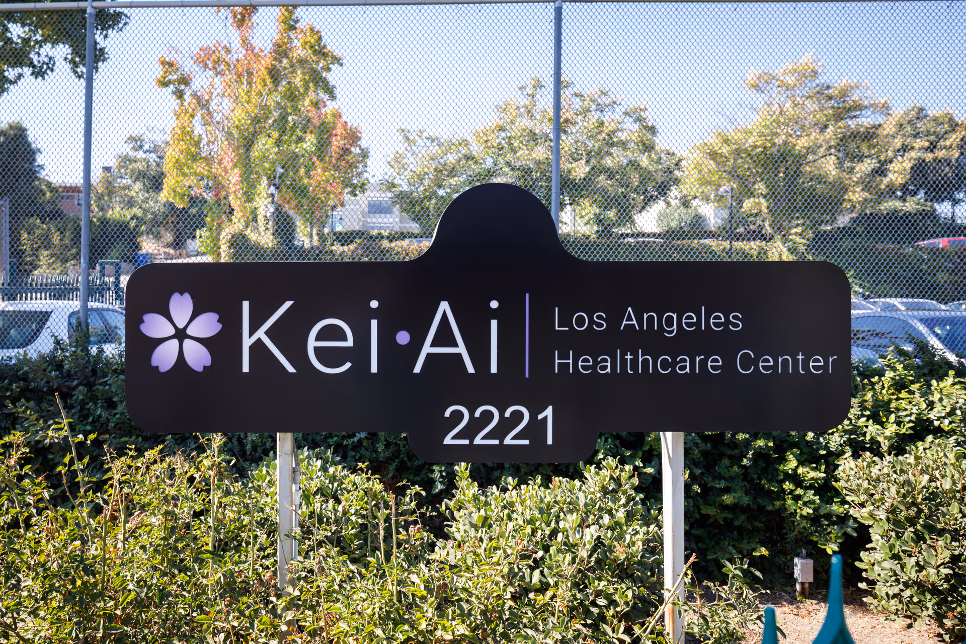Outdoor sign for Kei-Ai Los Angeles Healthcare Center with the address number 2221, surrounded by greenery and a chain-link fence in the background.