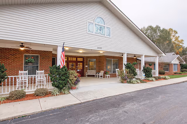 Front exterior view of Brookdale Hickory Northeast facility showing a covered entrance with white rocking chairs, potted plants, an American flag, and a brick facade under a gabled roof.