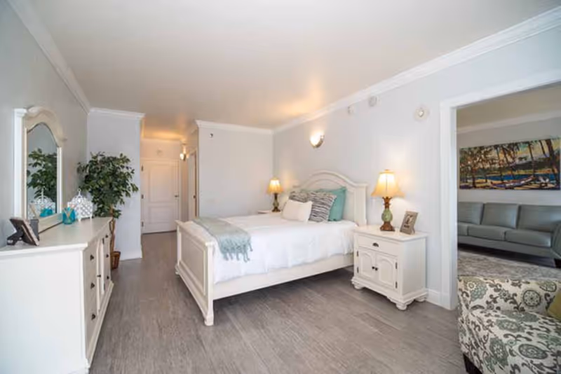 A bright and clean bedroom with a white bed frame, white bedding, and decorative pillows. There are two bedside tables with lamps on each side of the bed. A white dresser with a mirror and decorative items is on the left side of the room. A potted plant is next to the dresser. The floor is light-colored wood or laminate. Through an open doorway, a living room with a sofa, armchair, and a large painting on the wall is visible.