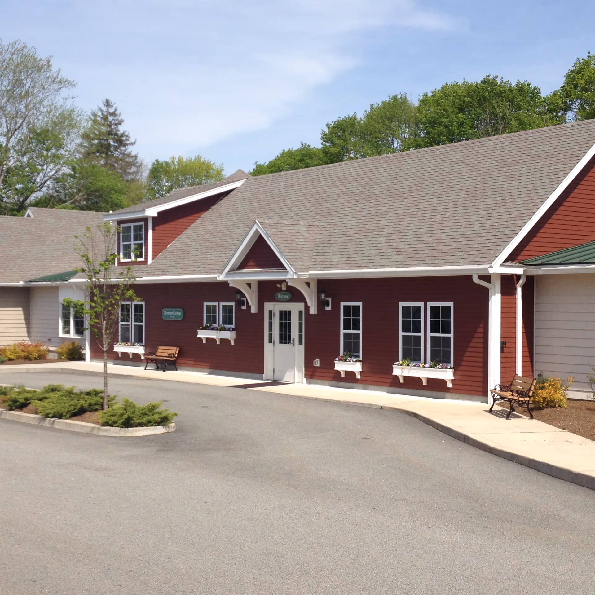 Exterior view of a red building with white trim and a gray roof, featuring multiple windows with flower boxes and two benches on a paved driveway. Trees and greenery are visible in the background under a partly cloudy sky.
