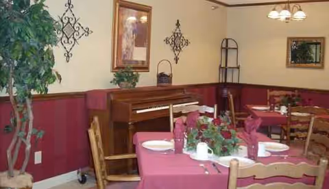 A dining room with red tablecloths, chairs, plants, and a piano along the wall.