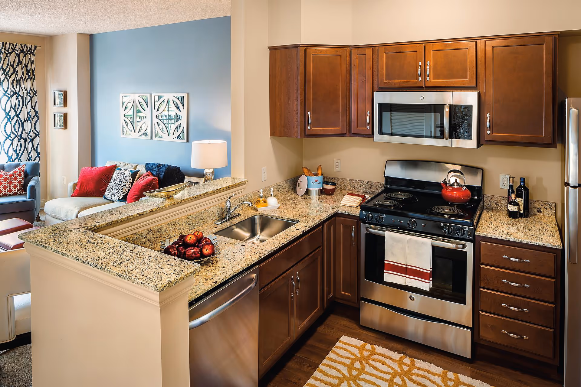 A modern kitchen with granite countertops, wooden cabinets, a stainless steel stove with a red kettle, a microwave, and a refrigerator. The kitchen opens into a living area with a beige sofa, red and patterned cushions, a blue accent wall, and a lamp on a side table.
