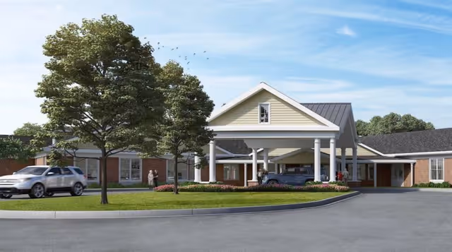 Exterior view of a senior living facility with a covered entrance supported by white columns. There are two cars parked near the entrance and a few people standing outside. The building has a brick facade with a light-colored gabled roof and is surrounded by green trees and landscaped grass areas under a blue sky.
