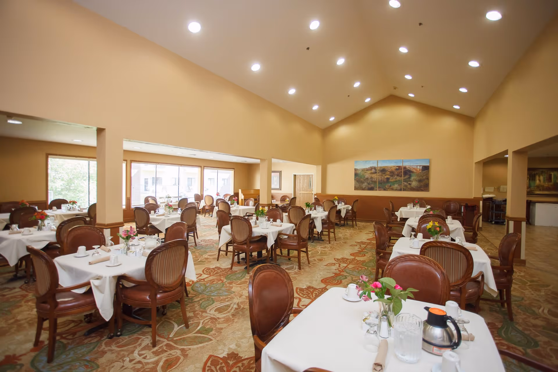 A spacious dining room with multiple tables covered in white tablecloths, each set with cups, glasses, napkins, and small flower arrangements. The room has high ceilings with recessed lighting, large windows letting in natural light, and a patterned carpet. There are wooden chairs with cushioned seats around the tables and a landscape painting on the far wall.