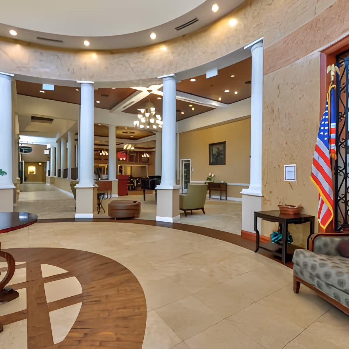 Interior view of a senior living facility lobby area with a circular floor design, white columns, beige walls, and ceiling lights. There is a seating area with chairs and tables, an American flag on the right side, and a hallway leading to other rooms.