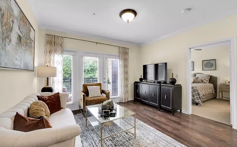 Bright living room with a white sofa, armchair, glass coffee table, TV on a black console, and a doorway to a bedroom.