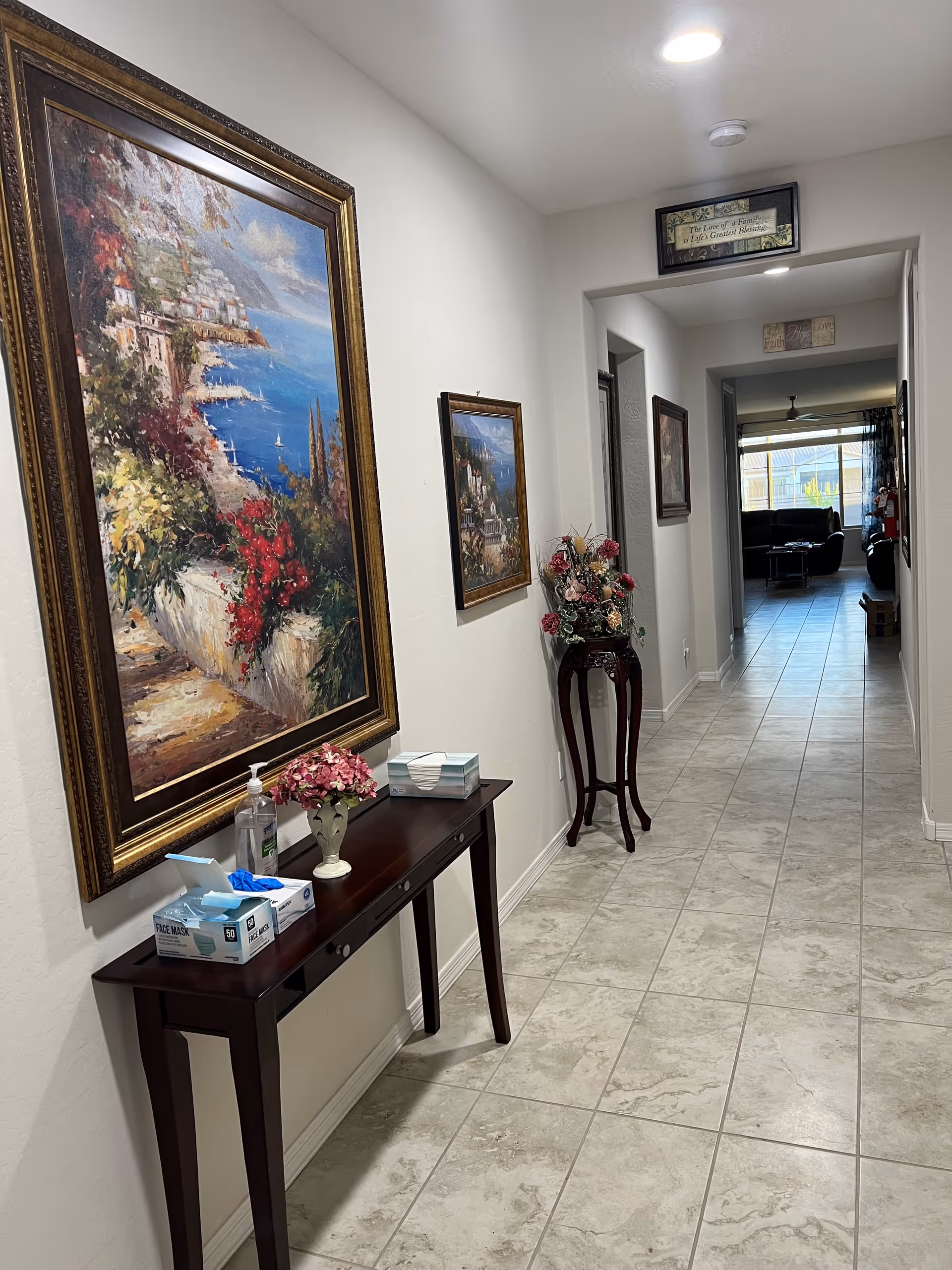 A bright tiled interior hallway lined with framed paintings, a small console table holding hand sanitizer and face masks, and a view into a living area at the end.