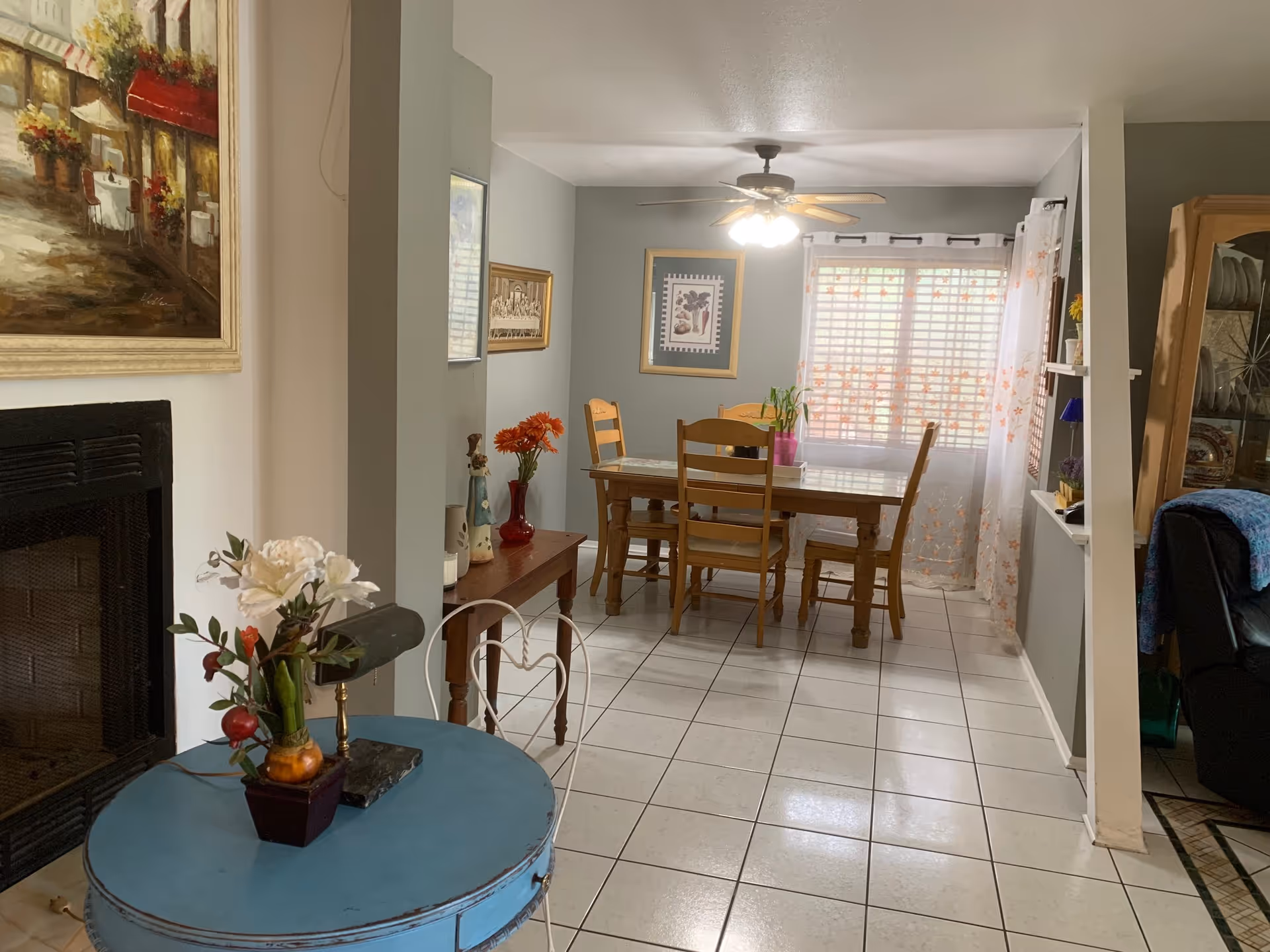 A cozy dining room with a wooden dining table and four chairs. The room has light gray walls, a ceiling fan with lights, and a window with white blinds and sheer curtains with orange floral patterns. There is a small blue round table with a flower arrangement in the foreground, a fireplace on the left, and framed artwork on the walls. A wooden cabinet with dishes is visible on the right side.
