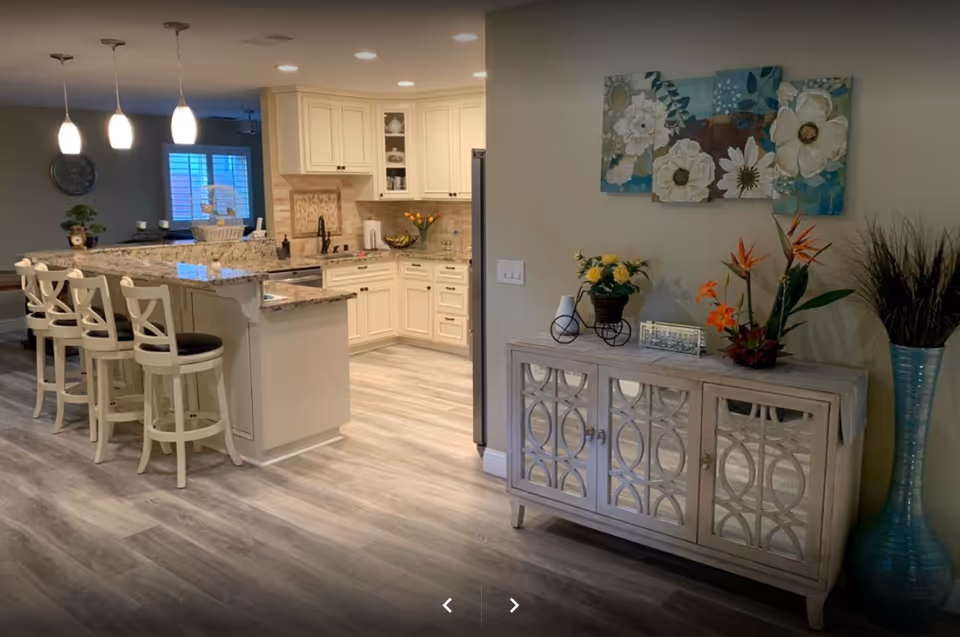 A modern kitchen and dining area with a granite countertop island and four white bar stools. The kitchen features cream-colored cabinets, a stainless steel refrigerator, and decorative items including flowers and a basket. Adjacent to the kitchen is a wooden sideboard with mirrored doors, decorated with flower arrangements and a floral painting on the wall above it. The flooring is light wood, and pendant lights hang above the kitchen island.