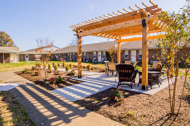 Outdoor patio area at Red Cedar Glen featuring a wooden pergola with seating underneath, surrounded by landscaped garden beds and a paved walkway, with single-story brick buildings in the background under a clear blue sky.
