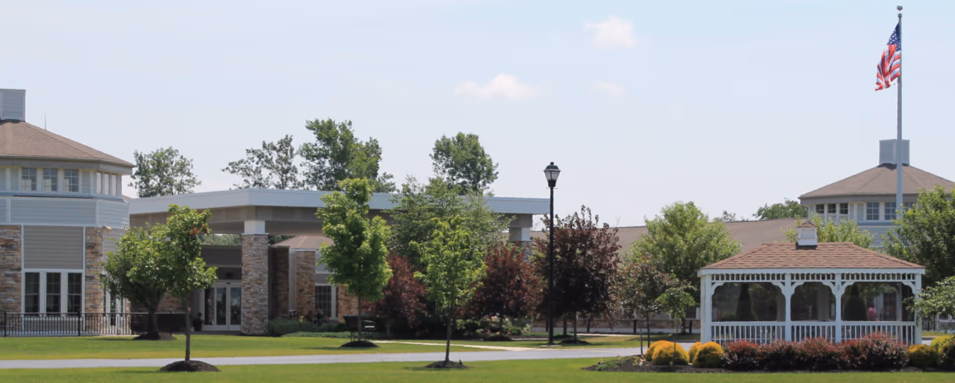 Exterior view of a senior living facility with a well-maintained lawn, several trees, a white gazebo, and an American flag on a flagpole. The building has stone and siding exterior with multiple windows and a covered entrance.