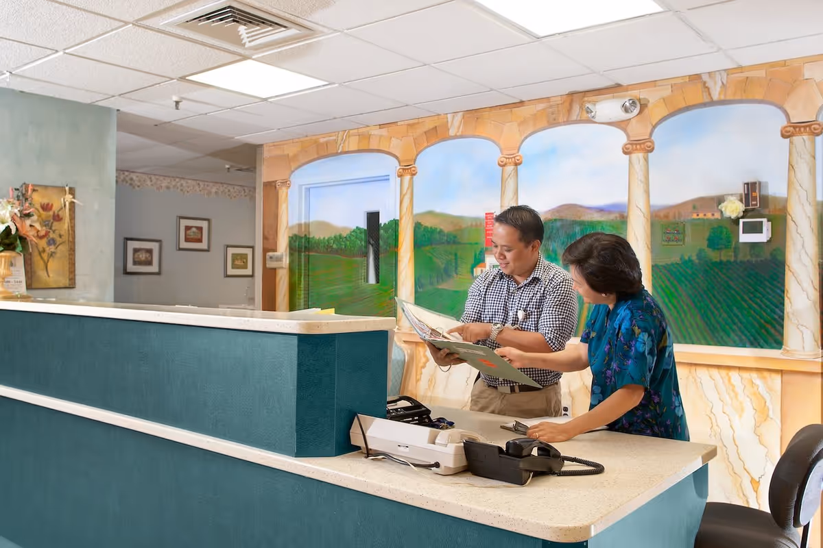 Two people standing behind a reception desk in a care facility, looking at a folder together. The wall behind them features a mural of a scenic landscape with columns and greenery.