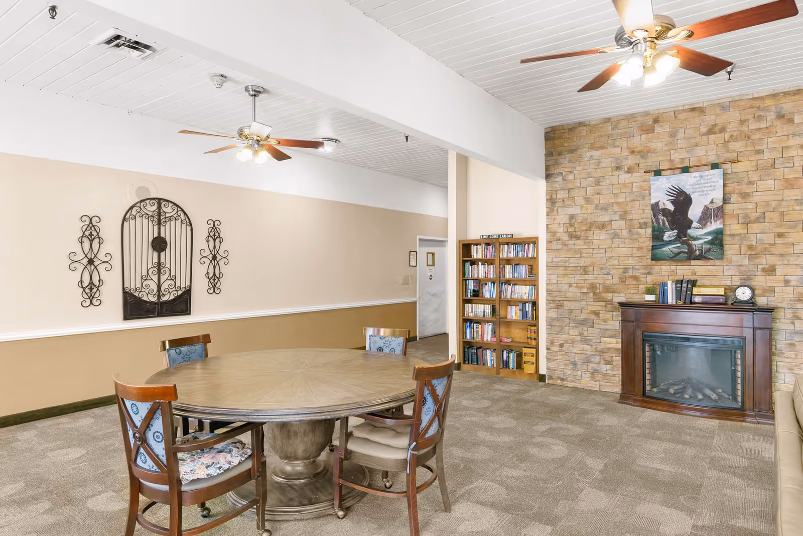 A cozy interior room with a round wooden table surrounded by four chairs with patterned cushions. The room features a stone accent wall with an electric fireplace below a painting of an eagle. A wooden bookshelf filled with books is next to the fireplace. The ceiling has white paneling with two ceiling fans with lights. The walls are painted beige with decorative metal wall art on one side.