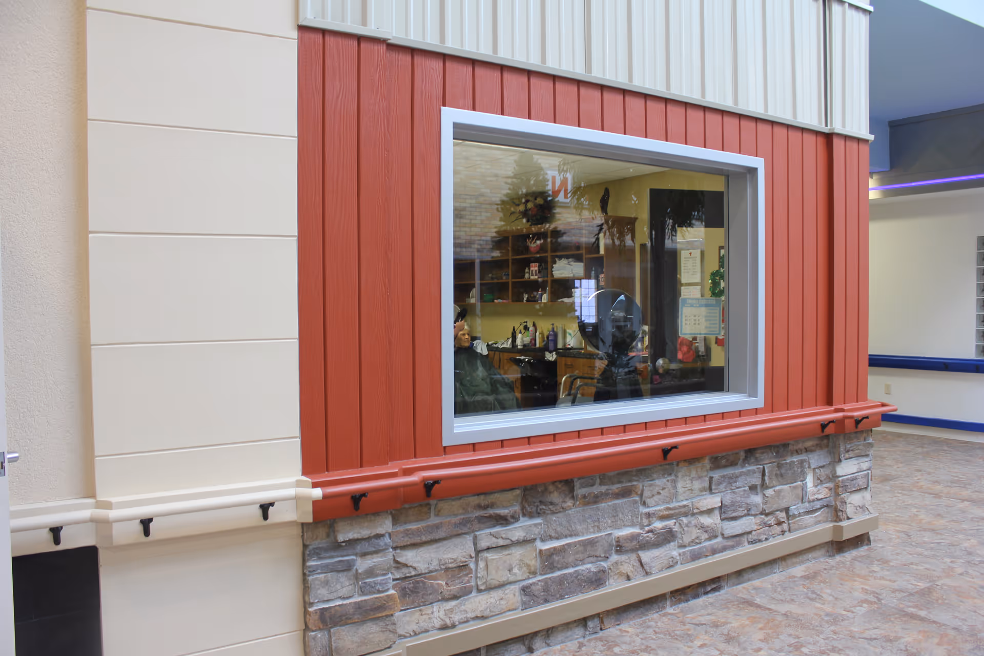 Interior view of a senior living facility showing a large window into a hair salon area where a person is seated under a hair dryer. The surrounding walls are decorated with beige, red vertical panels, and stone accents. The floor is tiled and the hallway outside the salon is visible.