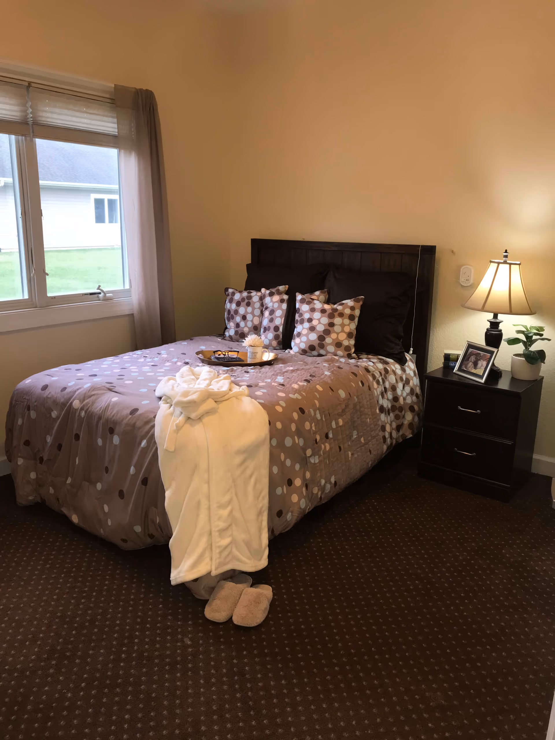 A cozy bedroom with a bed covered in a brown and beige polka dot comforter and matching pillows. A white robe and slippers are placed at the foot of the bed. There is a nightstand with a lamp, a small plant, and a framed photo. A window with blinds and sheer curtains lets in natural light.