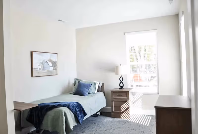 A bright bedroom with a single bed covered in green bedding and a blue throw blanket. There is a wooden nightstand with a lamp next to the bed, a window with blinds letting in sunlight, a framed picture on the wall, and a wooden dresser on the right side.