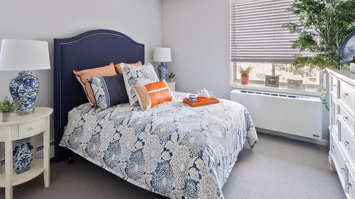 Bright, well-decorated bedroom with a navy upholstered headboard, patterned bedding, bedside tables with lamps, and a dresser by a window.