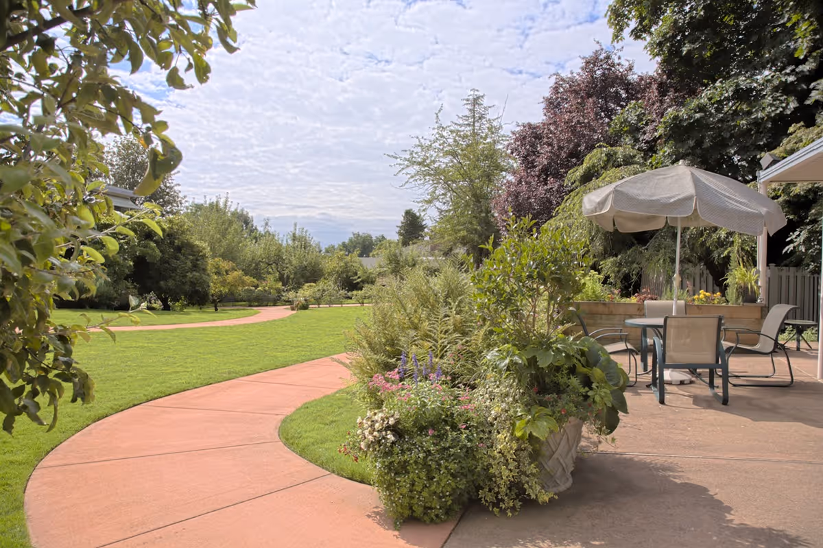 A curved paved walkway through a green garden area with various trees and shrubs. On the right side, there is a patio with a table, chairs, and a large umbrella providing shade. The sky is partly cloudy.