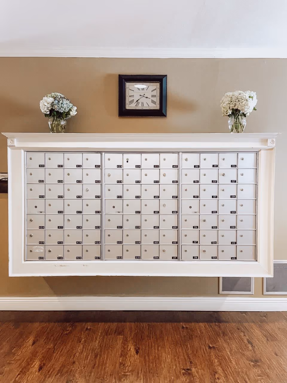 A wall-mounted white mailbox unit with multiple small locked compartments, each labeled with numbers. Above the mailbox unit is a square wall clock with Roman numerals. On top of the mailbox unit, there are two glass vases with white and light blue flowers. The wall behind is painted beige, and the floor is wooden.