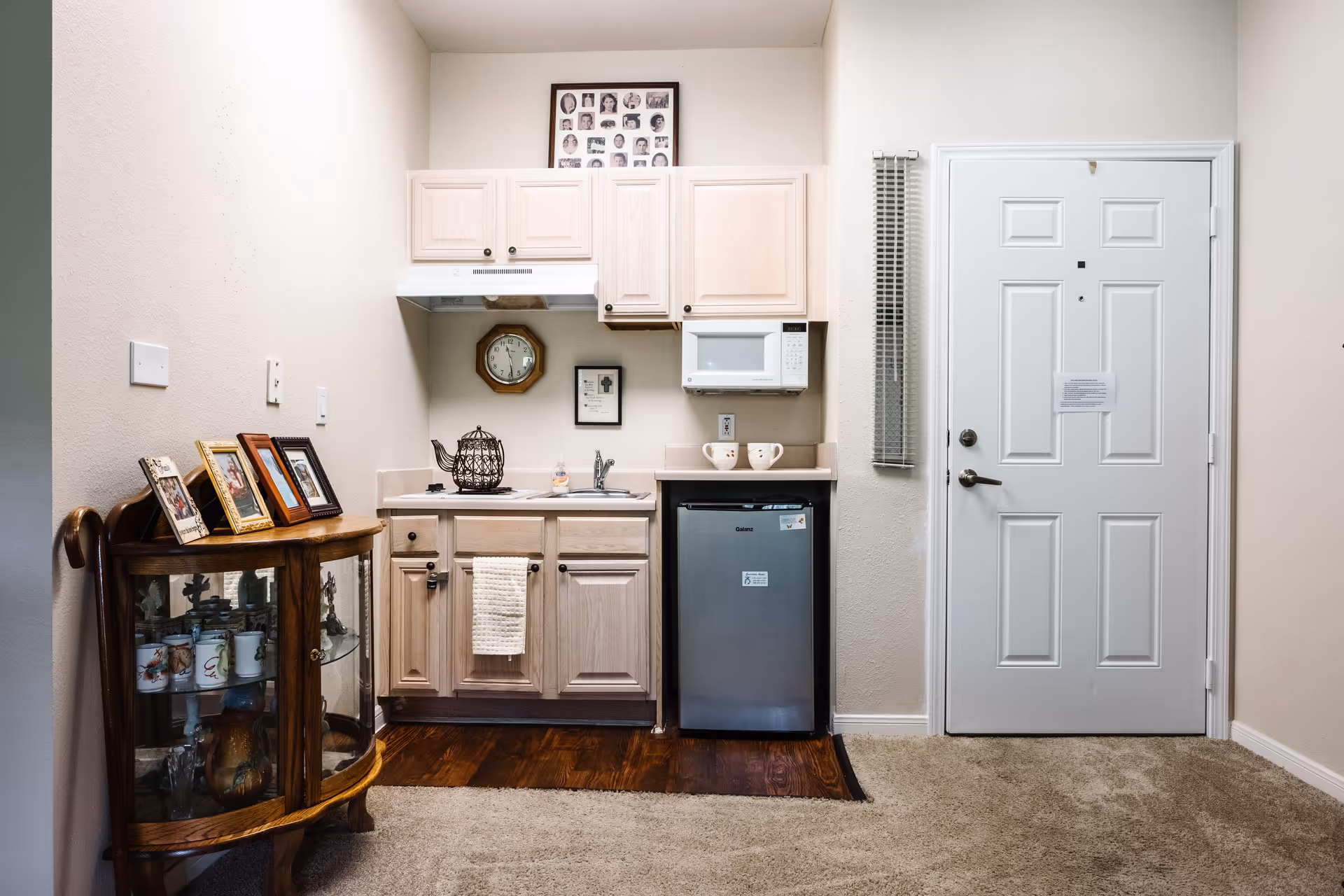 Small kitchenette area with light wood cabinets, a mini refrigerator, microwave, and a sink. There is a white door to the right, a small wooden display cabinet with framed photos on top to the left, and a clock and framed picture on the wall above the sink.