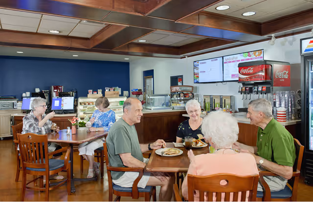 A group of elderly people sitting at wooden tables in a cafeteria-style dining area, enjoying food and drinks. The background shows a beverage dispenser with Coca-Cola branding, a refrigerated display case, and menu boards on the wall.