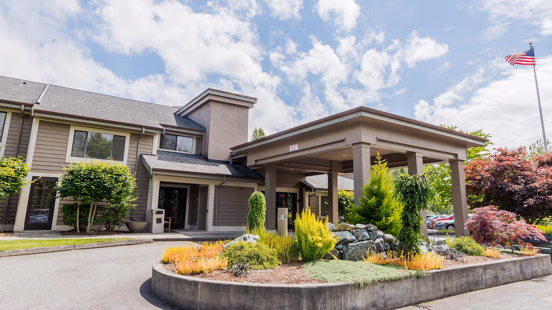 Exterior view of a senior living facility building with a covered entrance, landscaped garden with various shrubs and plants, a driveway, and an American flag on a flagpole under a partly cloudy sky.