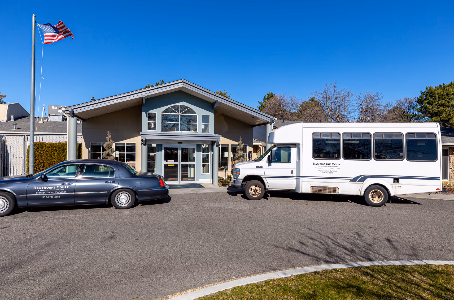 Front exterior view of Hawthorne Court facility with a clear blue sky. Two vehicles, a black sedan and a white shuttle bus, both labeled with Hawthorne Court Transportation Services, are parked in front of the entrance. An American flag is flying on a flagpole to the left of the building.