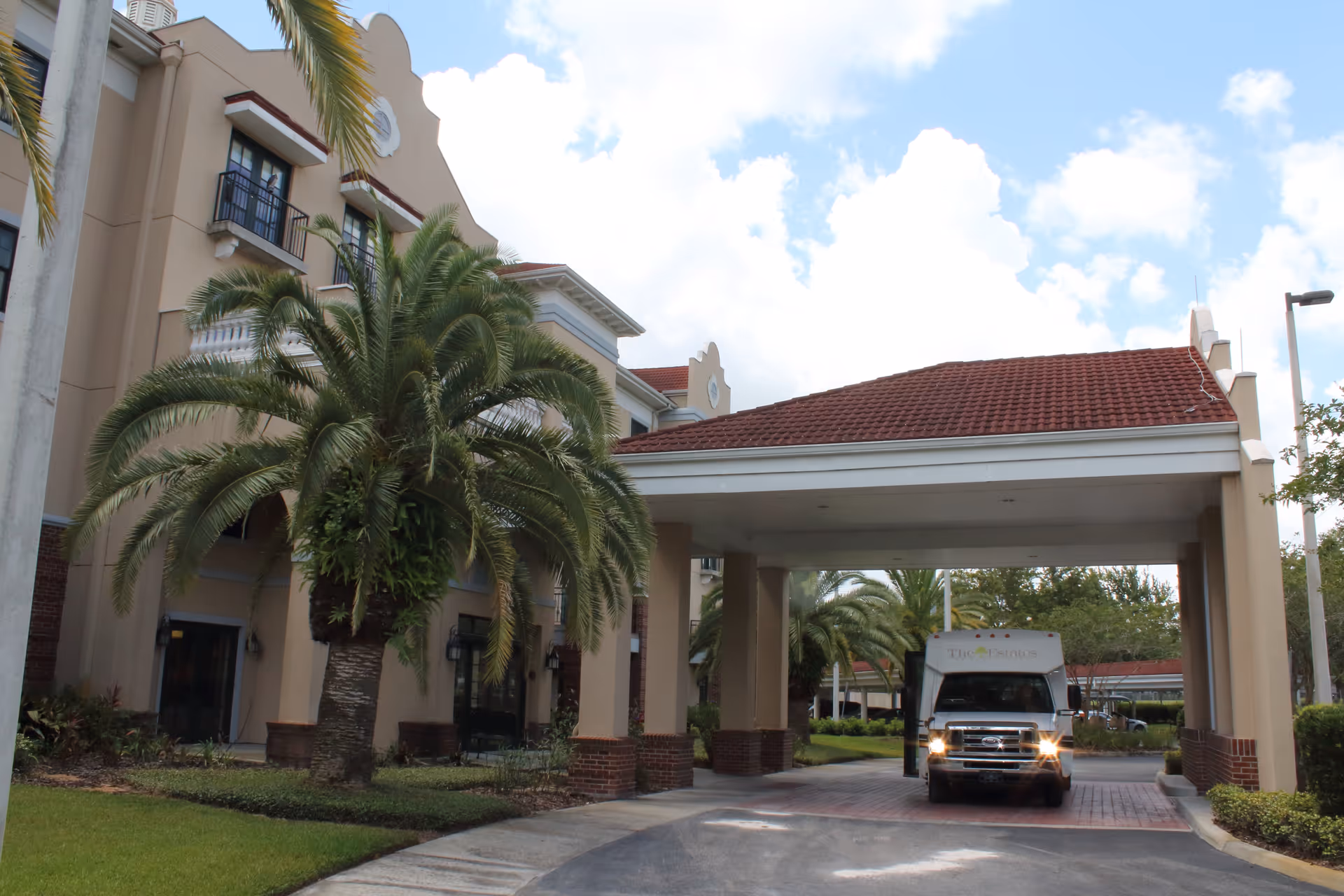 Entrance of a Mediterranean-style senior living building with a covered porte-cochère, palm trees, and a shuttle van.
