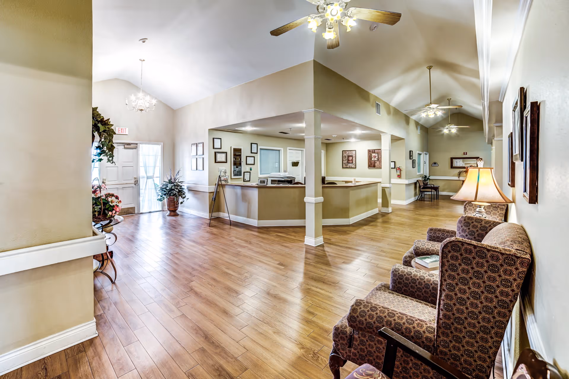 Interior view of a senior living facility lobby with wooden flooring, beige walls, and a reception desk in the center. There are patterned armchairs with a lamp on the right side, framed pictures on the walls, ceiling fans with lights, and potted plants near the exit door.
