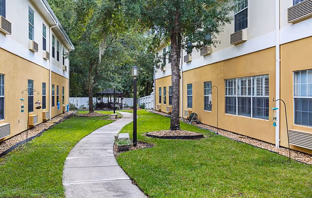 A paved walkway curves through a grassy courtyard between two beige two-story buildings with trees and a gazebo.