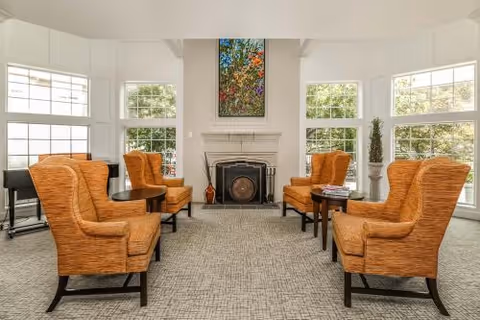 A bright and spacious living room with six orange upholstered armchairs arranged in pairs around small dark wooden tables. The room features large windows letting in natural light, a white brick fireplace with a colorful painting above it, and a light gray carpeted floor.