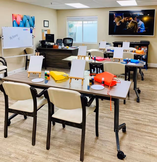 A bright activity room with tables and chairs arranged for a painting session. Each table has small easels with blank canvases, paint bottles, brushes, and colorful aprons. There is a whiteboard with a welcome message, a desk with a computer, and a large TV mounted on the wall showing a scene from a movie or show. The room has wood-style flooring and a window letting in natural light.