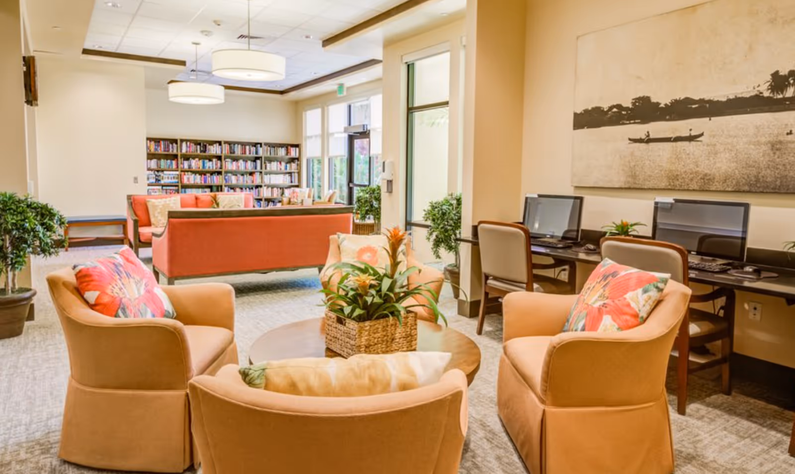 A bright and inviting common area with four beige armchairs arranged around a round wooden table with a plant centerpiece. In the background, there are two coral-colored sofas facing a bookshelf filled with books. To the right, there is a computer workstation with two desktop computers and chairs. Large windows let in natural light, and a large black and white wall art depicting a boat on water hangs above the computers.