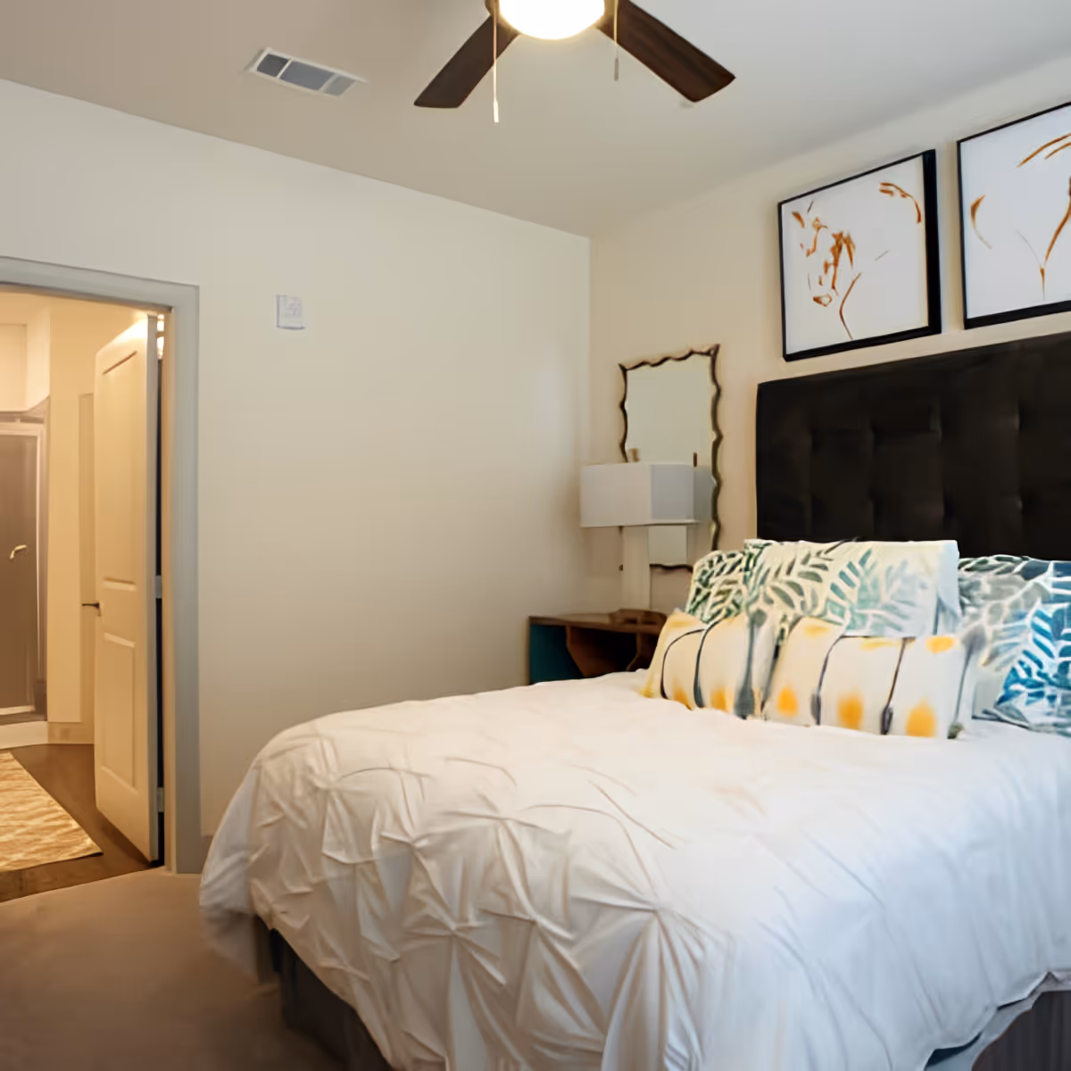 A bedroom with a neatly made bed featuring a white textured comforter and multiple decorative pillows with leaf patterns. Above the bed are two framed abstract art pieces. A ceiling fan with dark blades is visible on the ceiling. To the left, an open door leads to a bathroom with a shower and a rug on the floor. A bedside table with a lamp and a mirror is next to the bed.