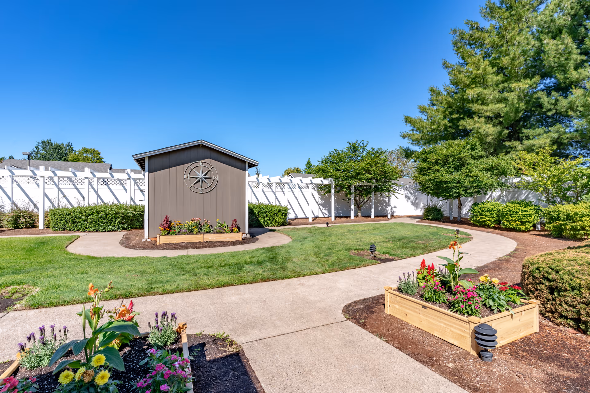 Well-maintained outdoor courtyard with winding concrete paths, raised flower beds, a small shed and a white fence.