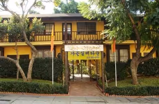 Front exterior view of a two-story building with yellow walls and brown trim, surrounded by trees and bushes. There is a gated entrance with a sign in Korean above it, and two flagpoles on either side of the entrance.