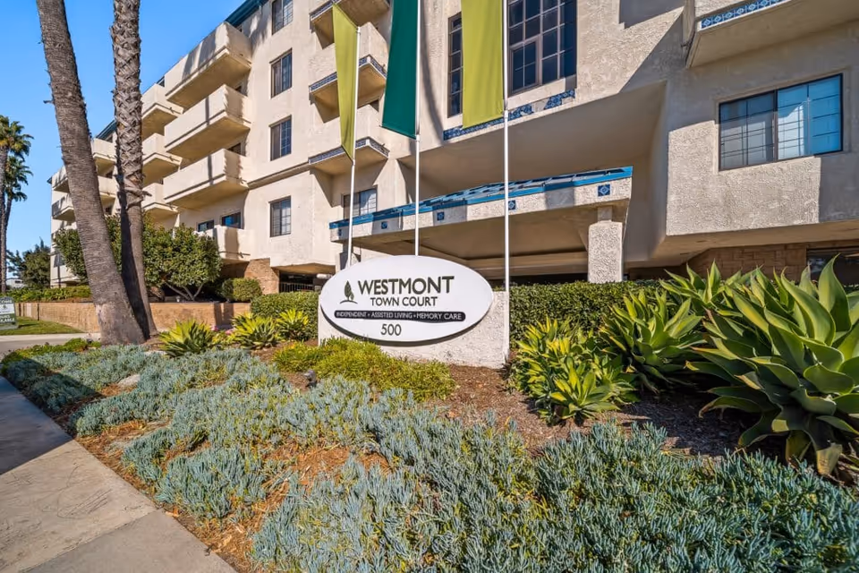 Exterior view of Westmont Town Court senior living facility with beige stucco building, balconies, green and yellow flags, and landscaped garden with succulents and palm trees.