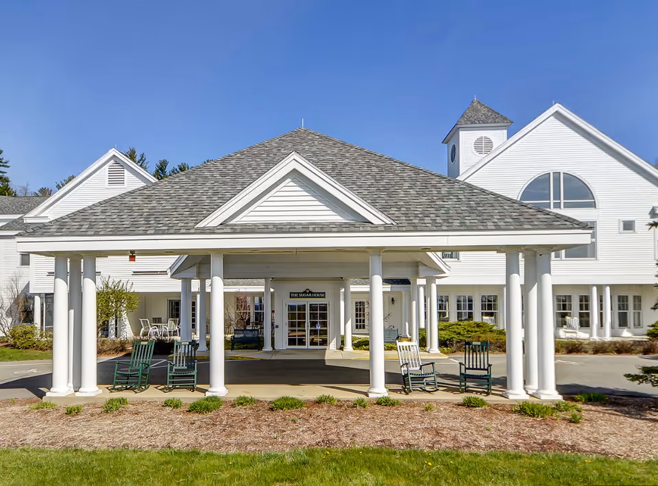 Front entrance of a white senior living building with a columned covered portico and rocking chairs on the driveway.