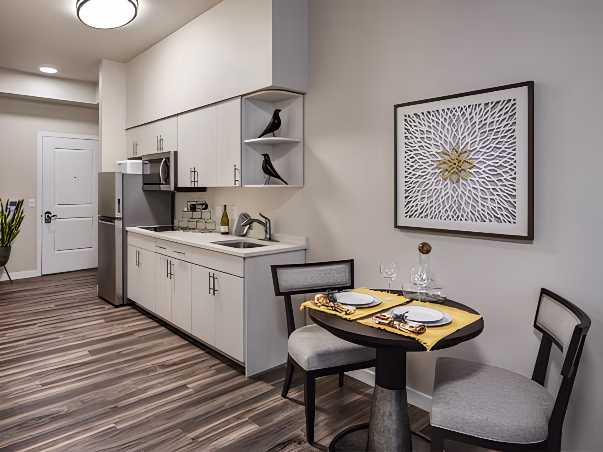 Small modern kitchenette with white cabinets, stainless refrigerator and sink, and a round two-person dining table set beneath wall art.
