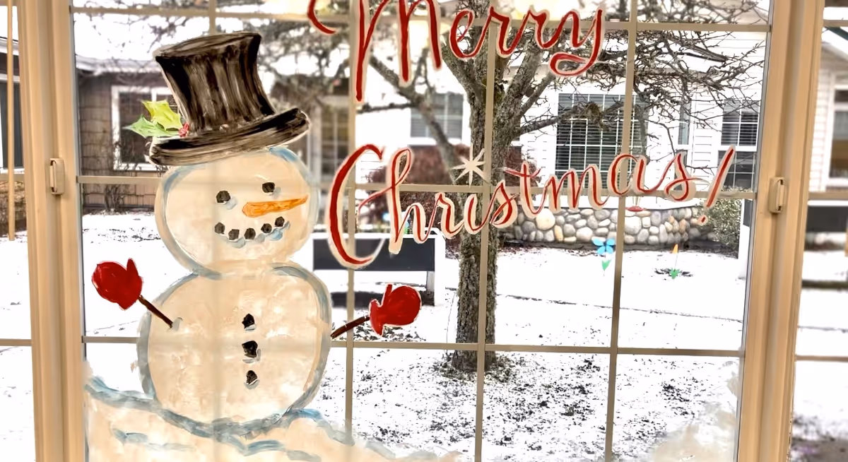 A window with a festive painting of a snowman wearing a black top hat and red mittens, with the words 'Merry Christmas!' written in red. Outside the window, there is a snowy yard with a tree and a building in the background.