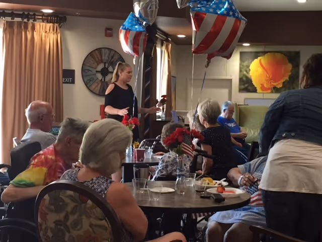 A group of elderly people seated around tables in a common area decorated with American flag-themed balloons. A woman is standing and interacting with the group. The room has warm lighting, a large clock on the wall, and a large flower painting in the background.