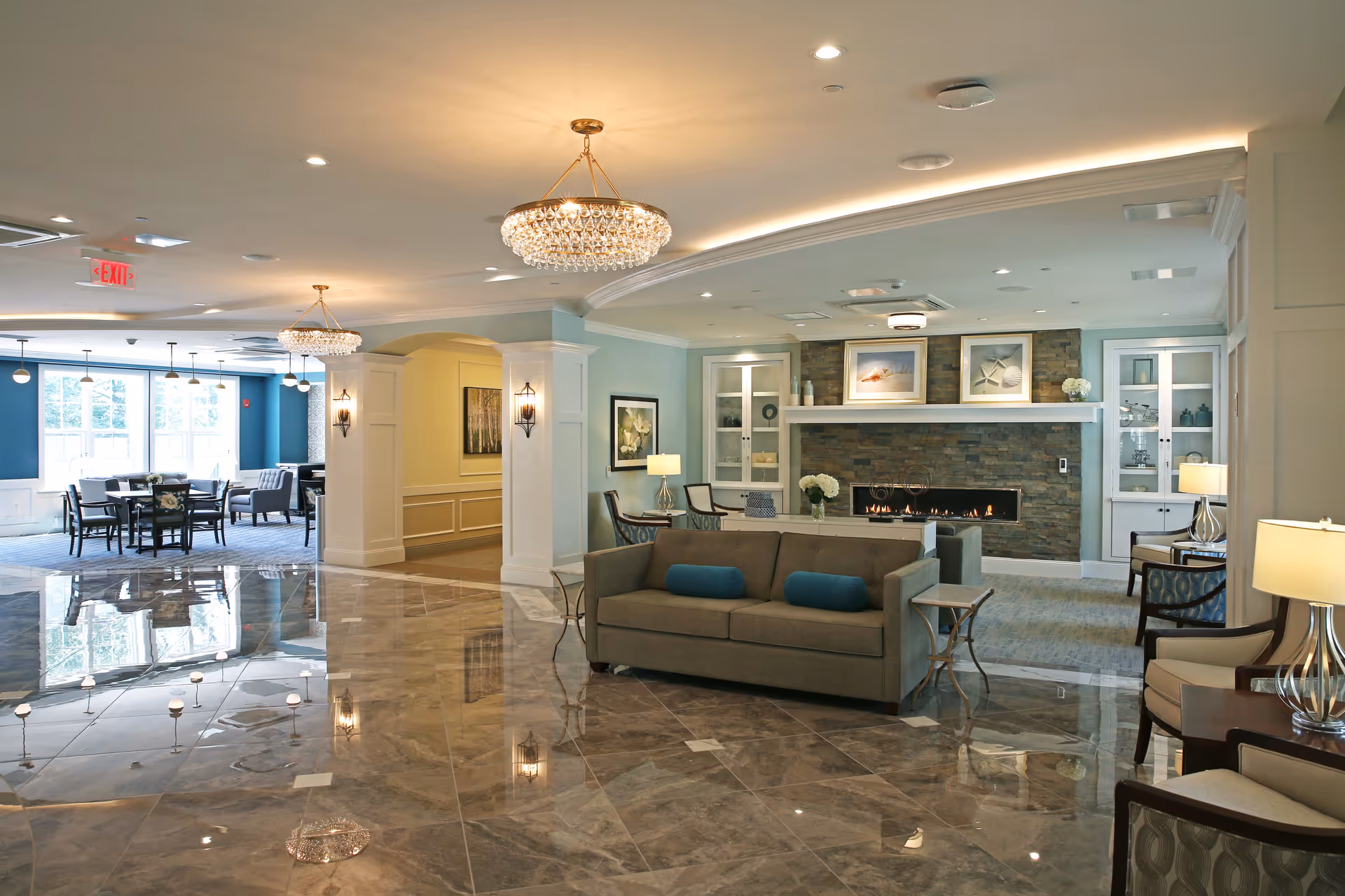 A spacious and elegant senior living common area featuring a polished marble floor, a gray sofa with blue cushions, armchairs, and side tables with lamps. The room includes a modern fireplace with a stone surround and decorative shelves on either side. In the background, there is a dining area with tables and chairs near large windows letting in natural light. The ceiling has recessed lighting and two crystal chandeliers.