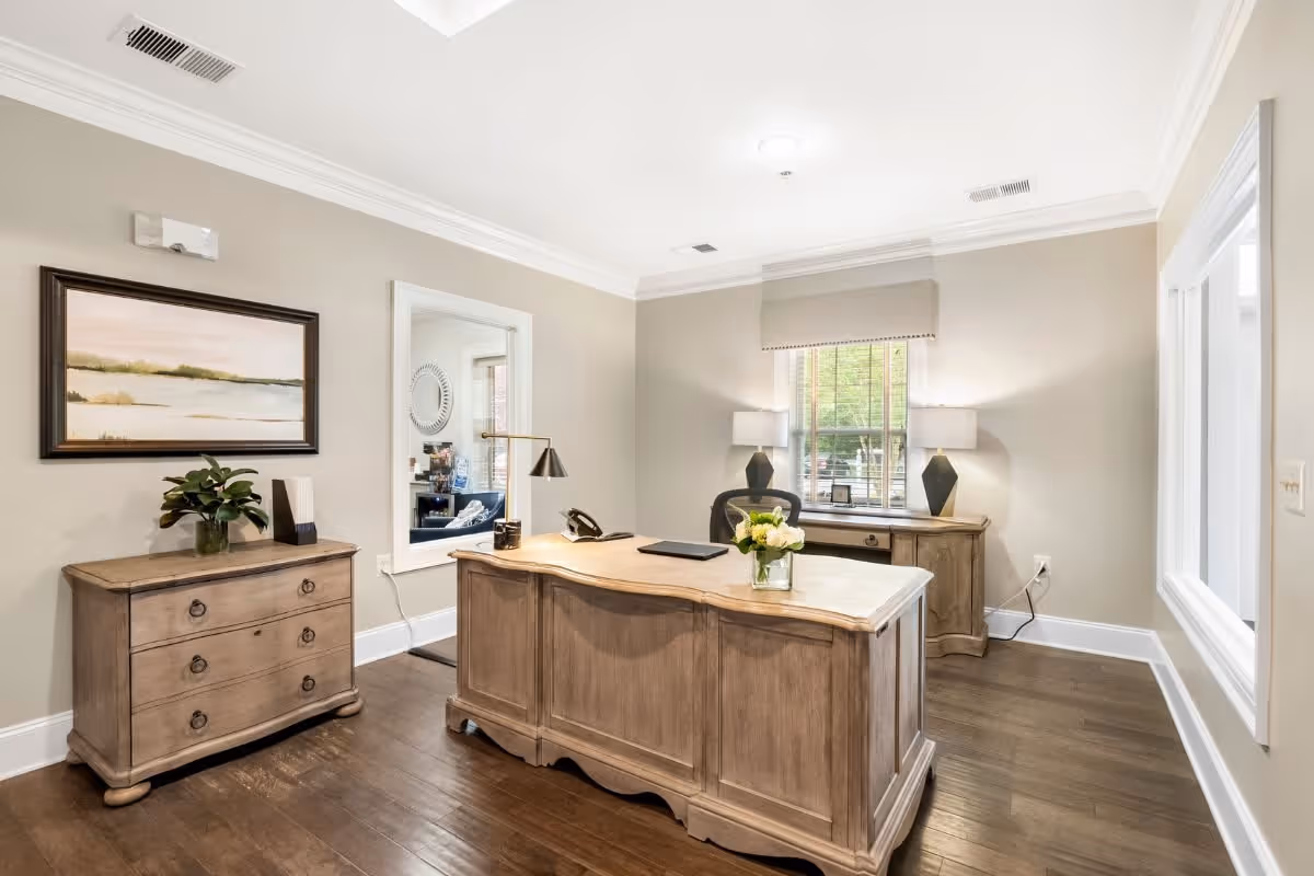 A bright office room with wooden flooring and beige walls. The room features a large wooden desk with a black office chair behind it, a small vase with flowers on the desk, and a desk lamp. There is a wooden dresser with a plant and decorative items on the left side, a window with a beige shade letting in natural light, and another wooden cabinet with two lamps on either side of the window.