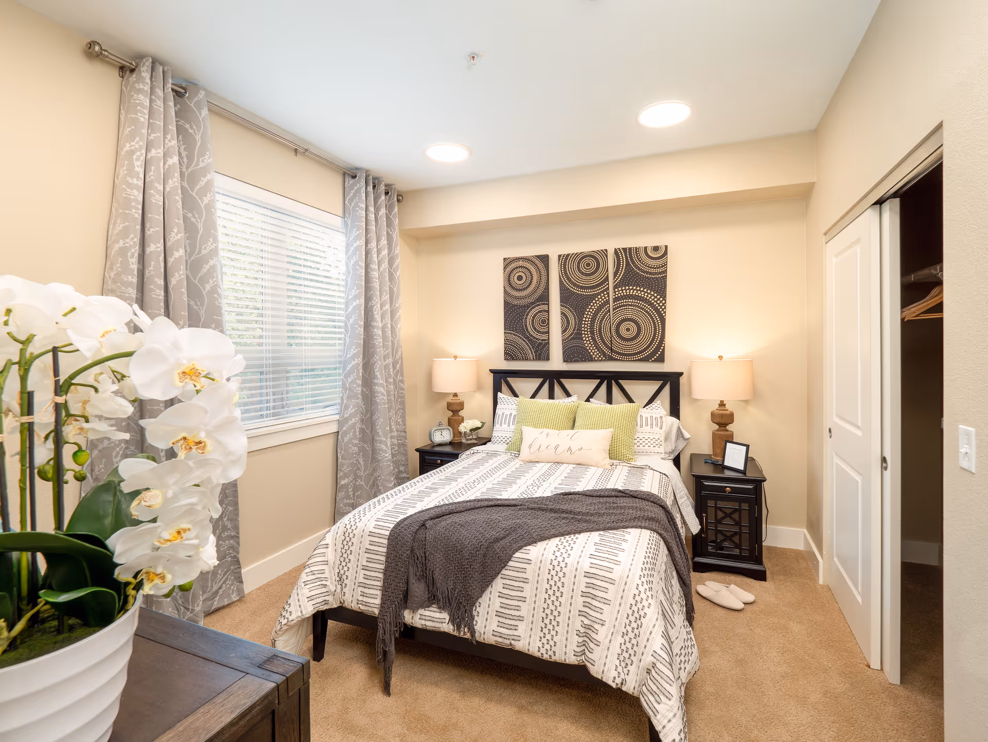 A cozy bedroom with a neatly made bed featuring patterned bedding and decorative pillows. Two bedside tables with lamps flank the bed. Above the bed are three pieces of circular patterned wall art. A window with gray patterned curtains is on the left wall, and a closet with sliding doors is on the right. A white orchid plant is visible in the foreground on a wooden dresser.