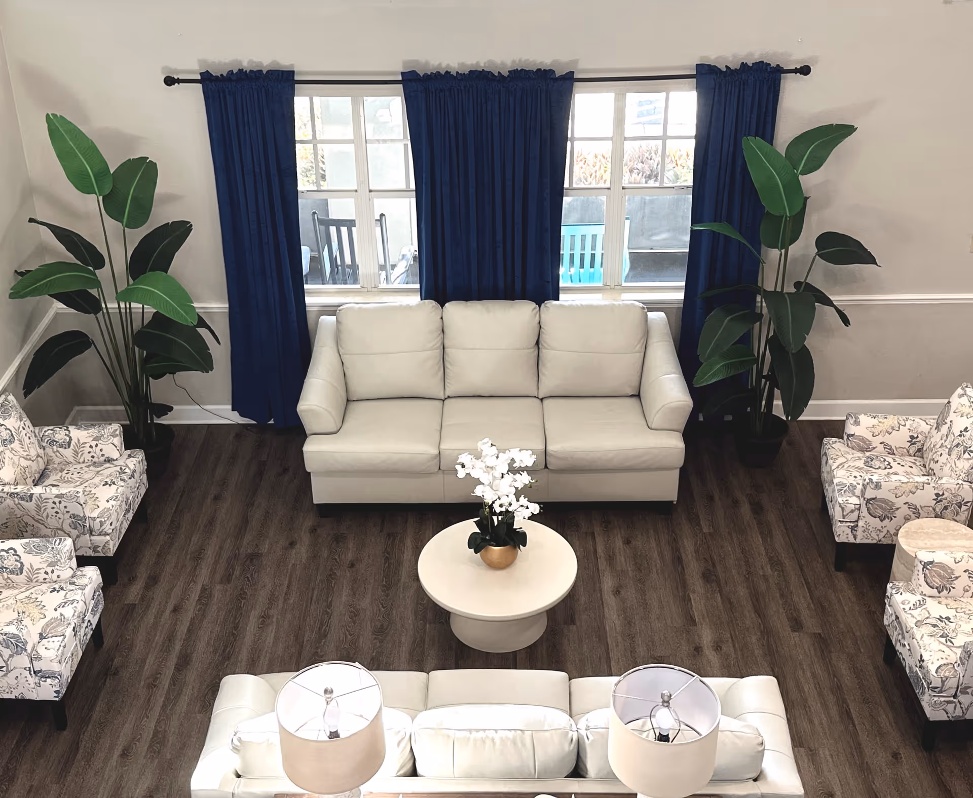 A bright living room with two white leather sofas facing each other, a round white coffee table with a white orchid plant in the center, two floral patterned armchairs on either side, two large green potted plants near the windows, and dark blue curtains covering three windows.
